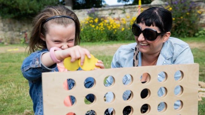 A mother and daughter are in a walled garden play a large wooden game of four in a row. The girl is dropping a yellow counter into the frame.